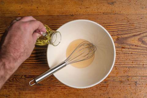 Hand gießt Öl in eine weiße Schüssel mit Schneebesen