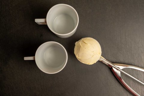 Eisportionierer aus Metall mit einer Eiskugel neben zwei weißen Tassen
