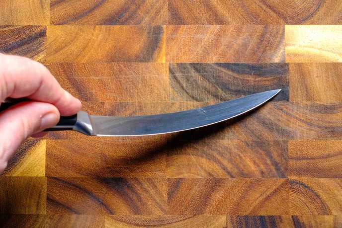 Hand hält ein Filetiermesser, dessen schmale Klinge sich über einem Holzbrett sichtbar biegt