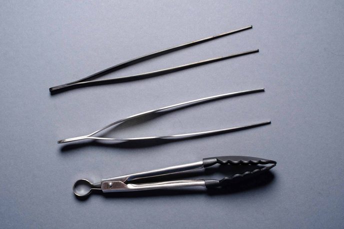 Zwei lange Pinzetten und eine Grillzange aus Edelstahl auf grauem Grund