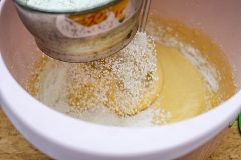 Mehl rieselt aus einem Bechersieb in hellen Rührteig