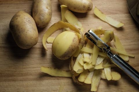 Rohe Kartoffeln und dünne Kartoffelschalen neben einem Pendelschäler auf dem Holzbrett