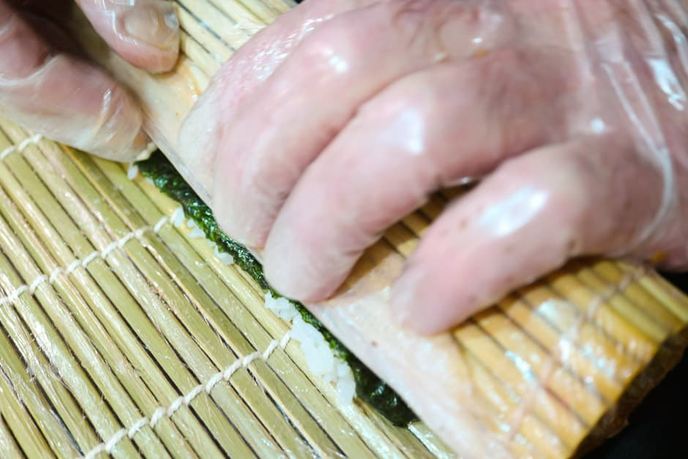 Hände in Handschuhen rollen Sushi mit einer Bambusmatte
